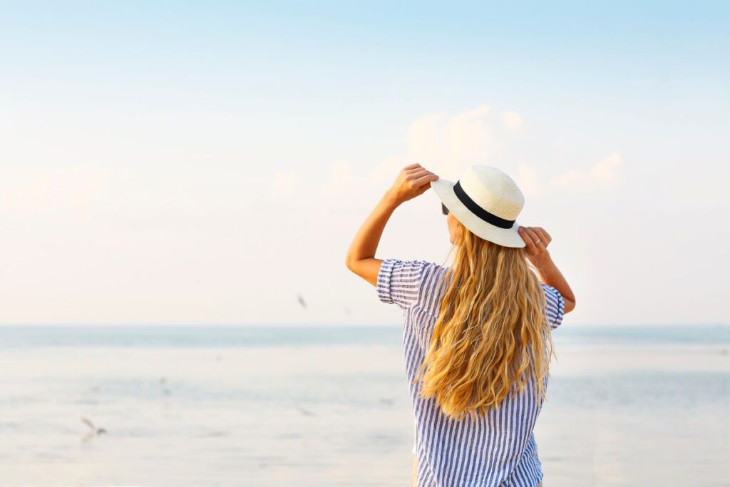 Blonde hair girl with a hat and a blue stripped shirt in front of the sea emphasizing how versatile a beach cover up can be in a Hawaii packing list.