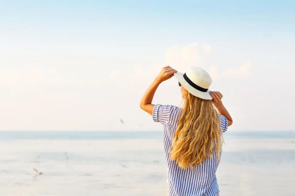 Blonde hair girl with a hat and a blue stripped shirt in front of the sea emphasizing how versatile a beach cover up can be in a Hawaii packing list.