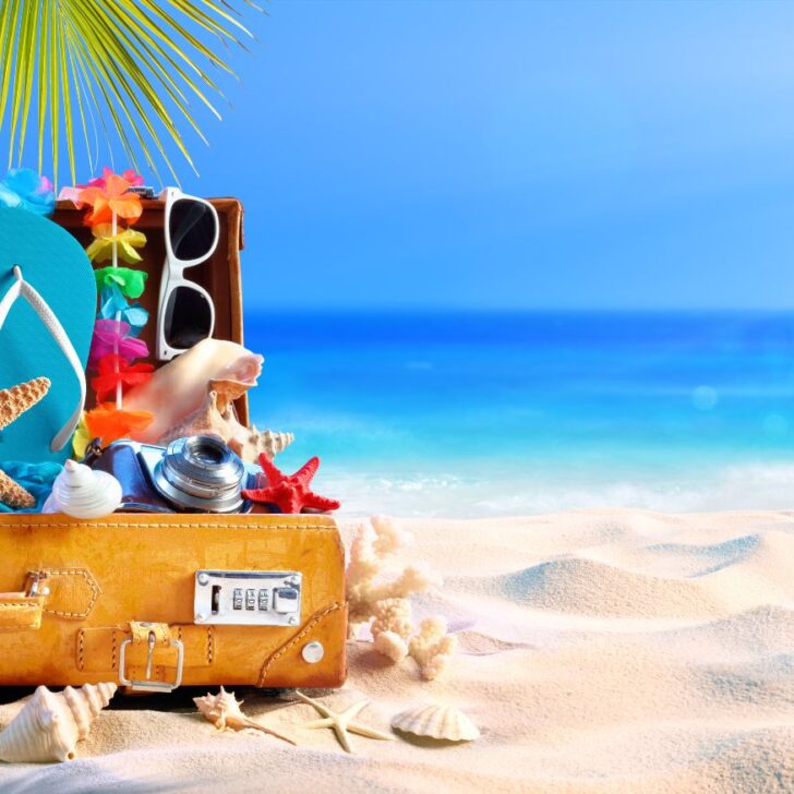 A suitcase half opened on a beach with all the essentials in a Hawaii packing list including sun glasses, flipflops, and more.