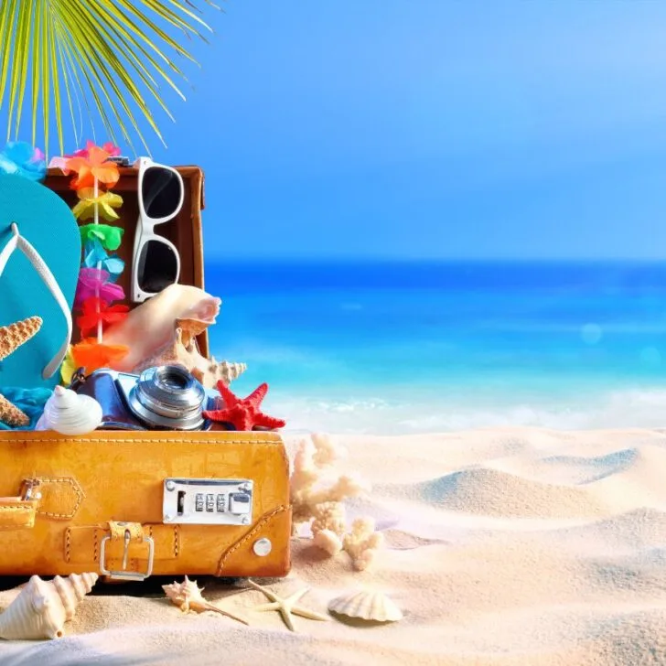 A suitcase half opened on a beach with all the essentials in a Hawaii packing list including sun glasses, flipflops, and more.