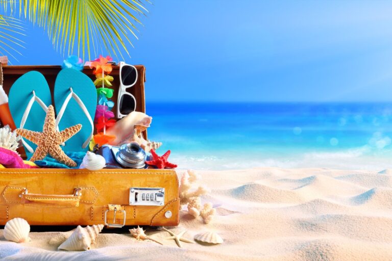A suitcase half opened on a beach with all the essentials in a Hawaii packing list including sun glasses, flipflops, and more.